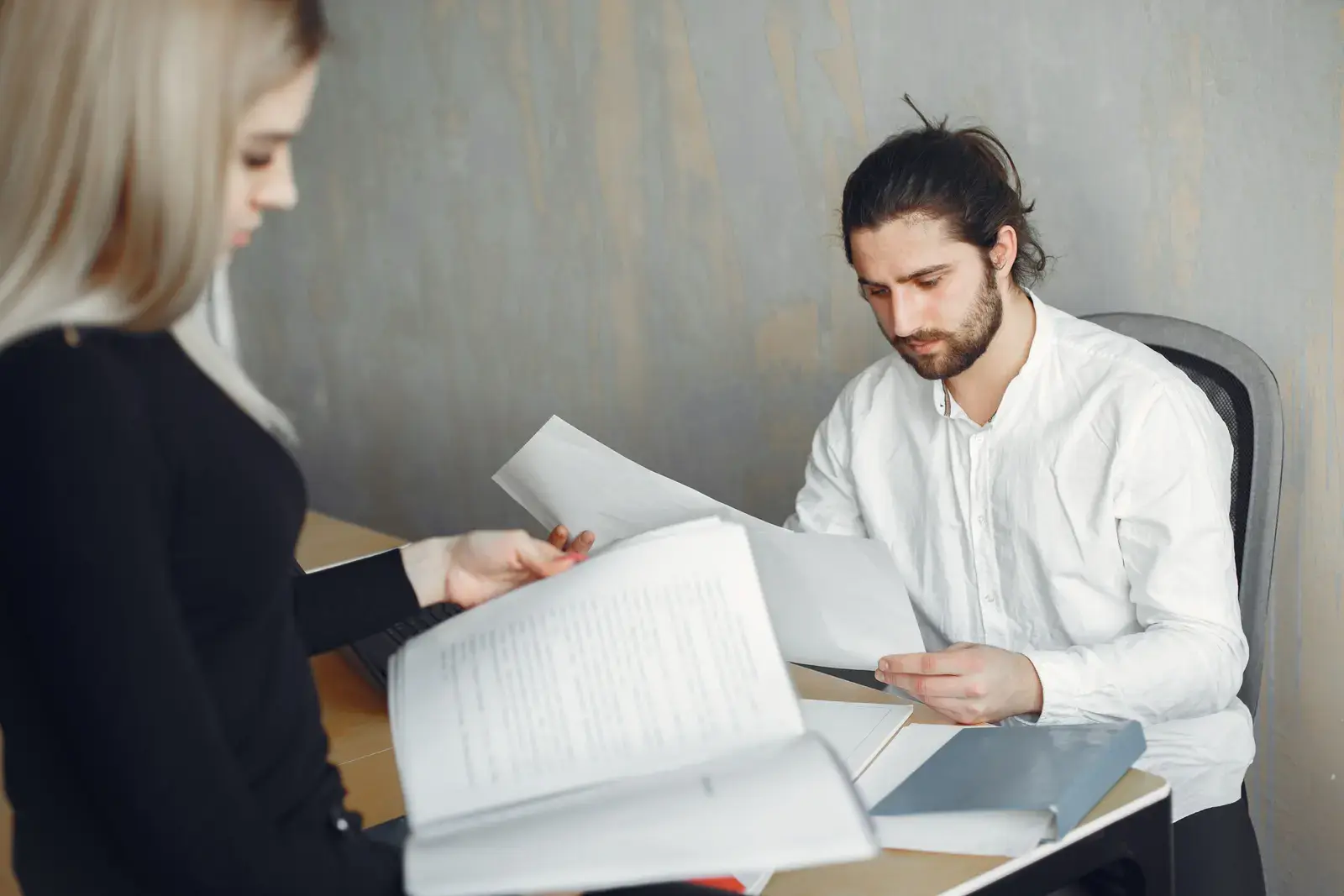 Two colleagues reading through printed documents and a bound report at a desk